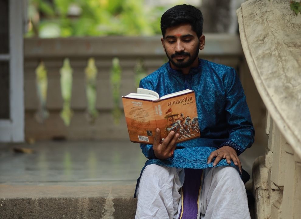 An image of a person sitting on a stone staircase and reading a book, with a background that relates to spirituality and culture. The person is wearing a blue kurta and white pants, which are traditional Indian garments. The book is orange in color and has a picture of a temple on it. The title of the book is “Mahabharata”, which is an ancient Indian epic that narrates the story of a war between two groups of cousins. The background consists of a porch with white pillars and greenery, creating a contrast with the stone staircase. The image conveys a sense of curiosity and learning.